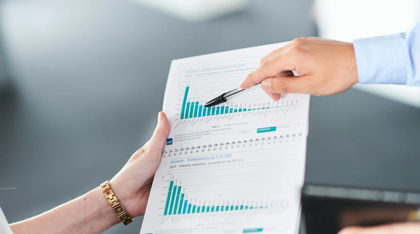 Two hands hold a printed report of teal bar charts while a pen points at the tallest bars.