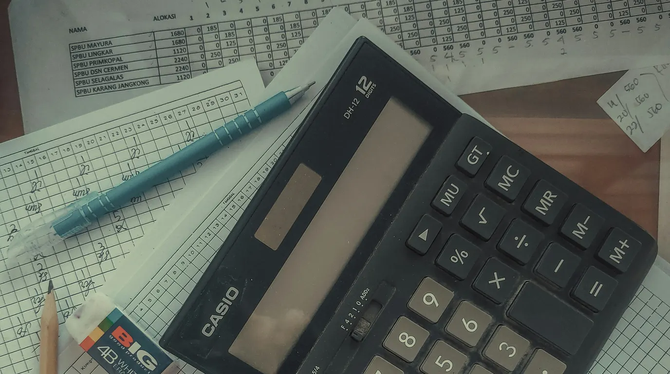 A large twelve-digit desk calculator lying across handwritten tally sheets, with a pencil, a slim pen and an eraser alongside.