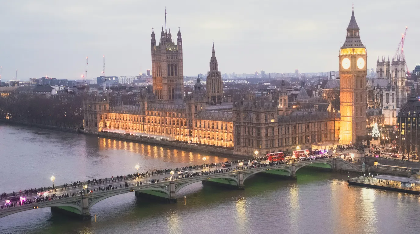 A floodlit gothic riverside palace and clock tower at dusk, with crowds crossing the arched bridge over the water below.