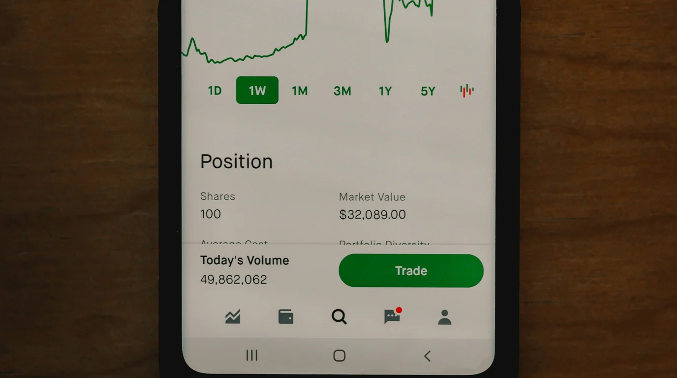 A phone on a dark wooden table showing a green trading screen with a share position, market value and a large trade button.