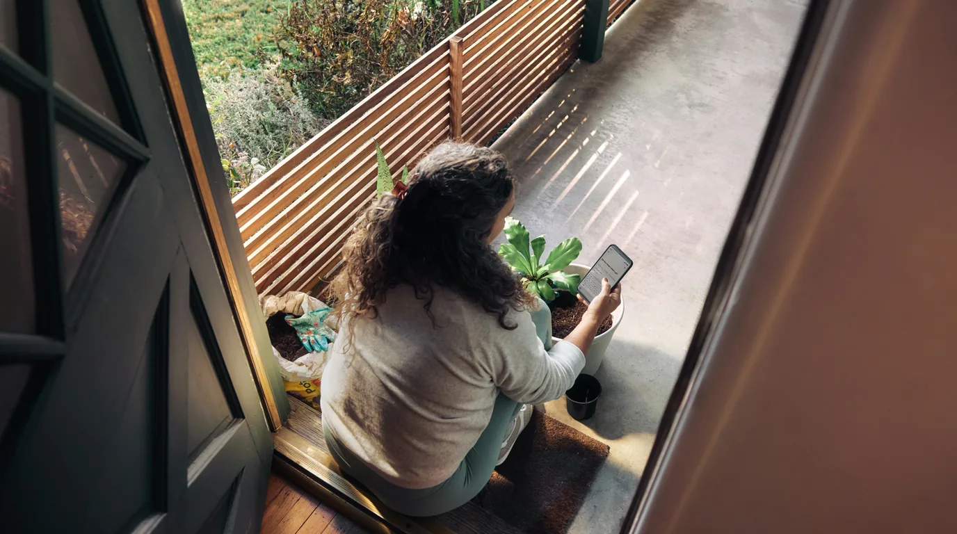 A woman sitting on a balcony bench in the sun, reading from a tablet.