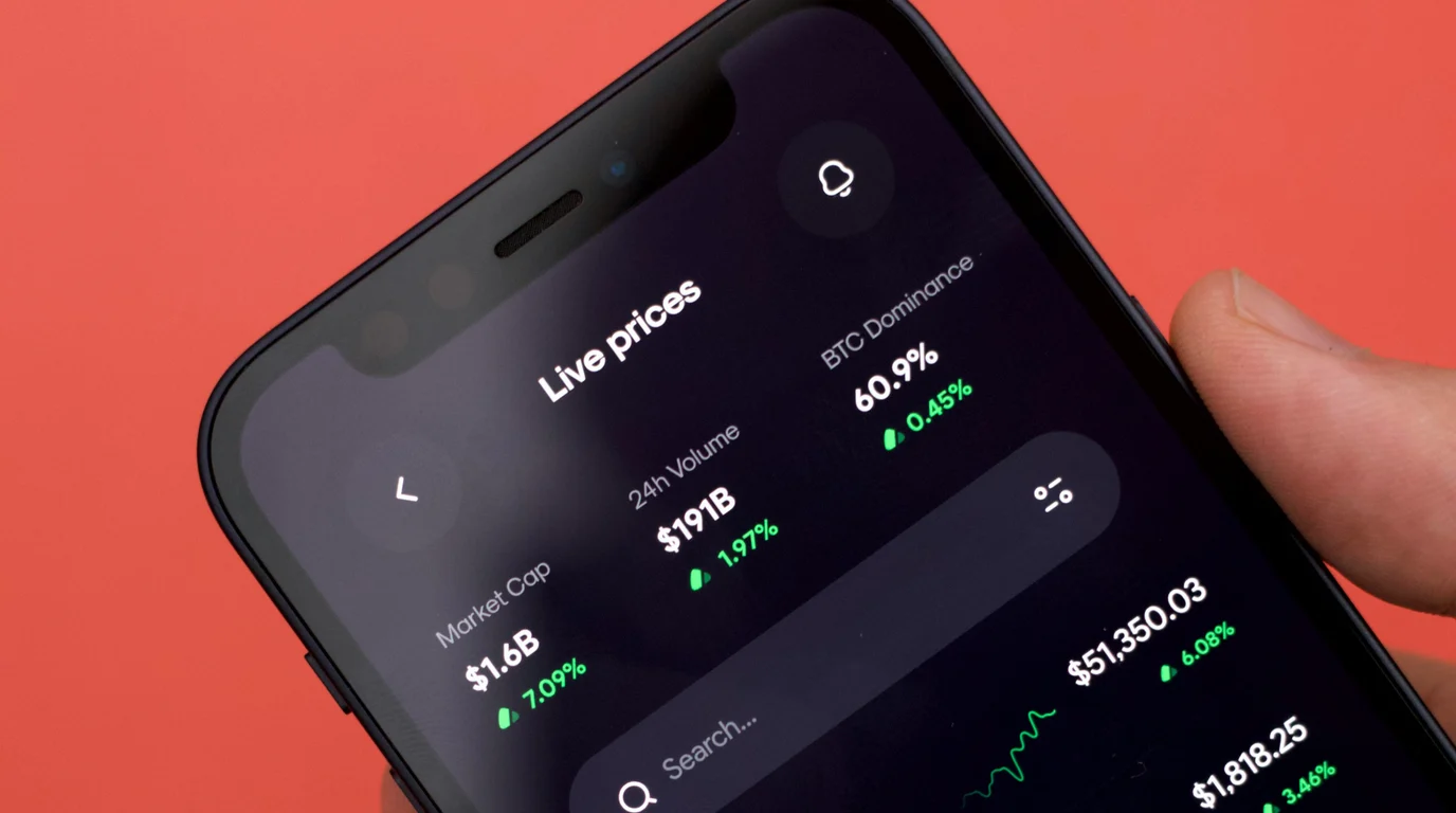 A phone held against a coral-red backdrop, its dark screen listing live market figures and green percentage changes.