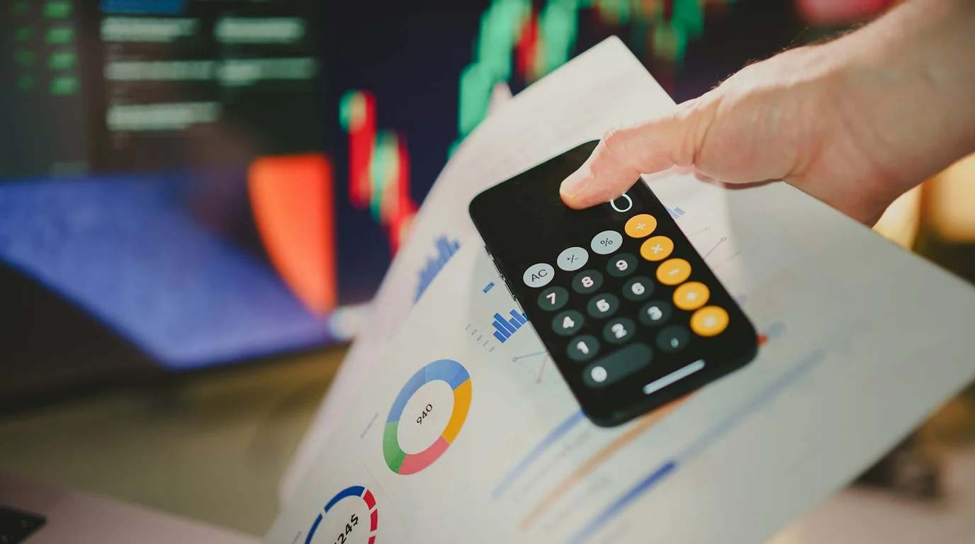 A hand tilts a phone open on its calculator app over printed pie and bar charts, with blurred candlestick screens behind.