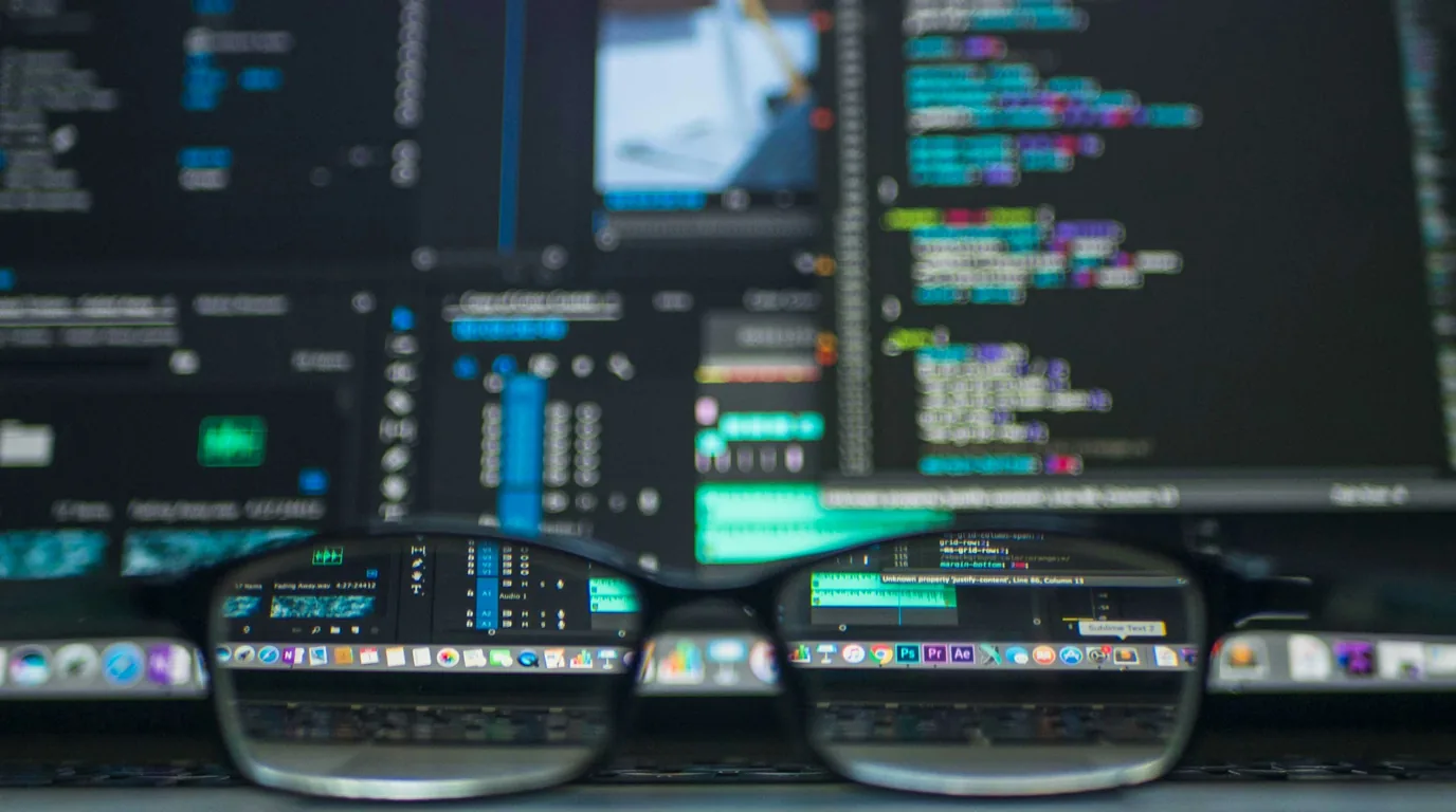 A pair of glasses standing on a desk, a screen of coloured code blurred behind them.