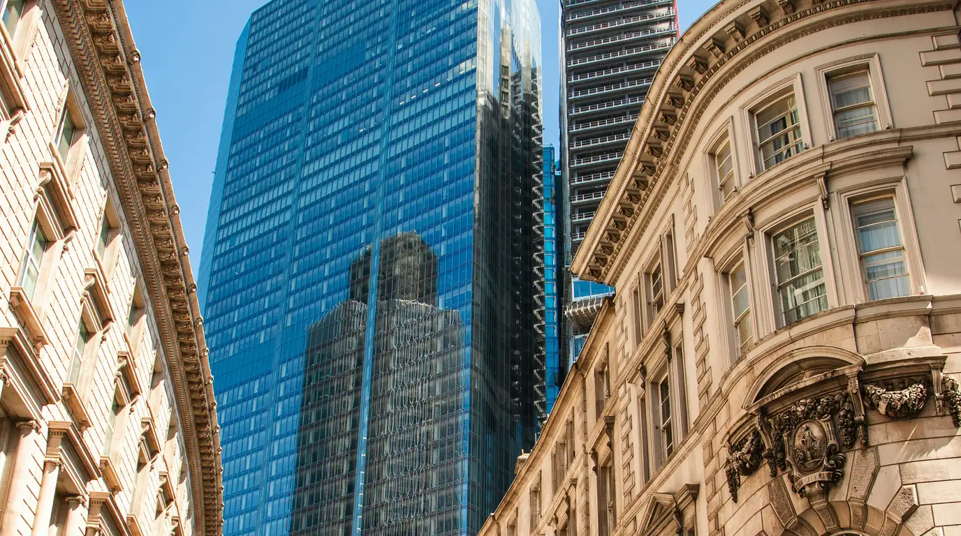 Looking up a narrow street between an ornate carved stone facade and a blue glass tower that mirrors its neighbours.