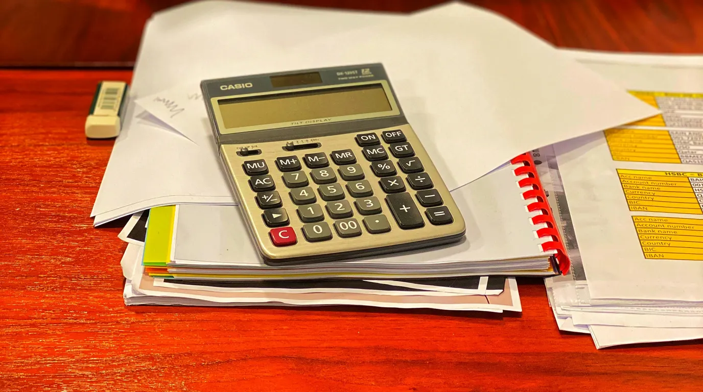 A desktop calculator resting on a stack of loose papers and a spiral-bound folder on a polished red-brown wooden desk.