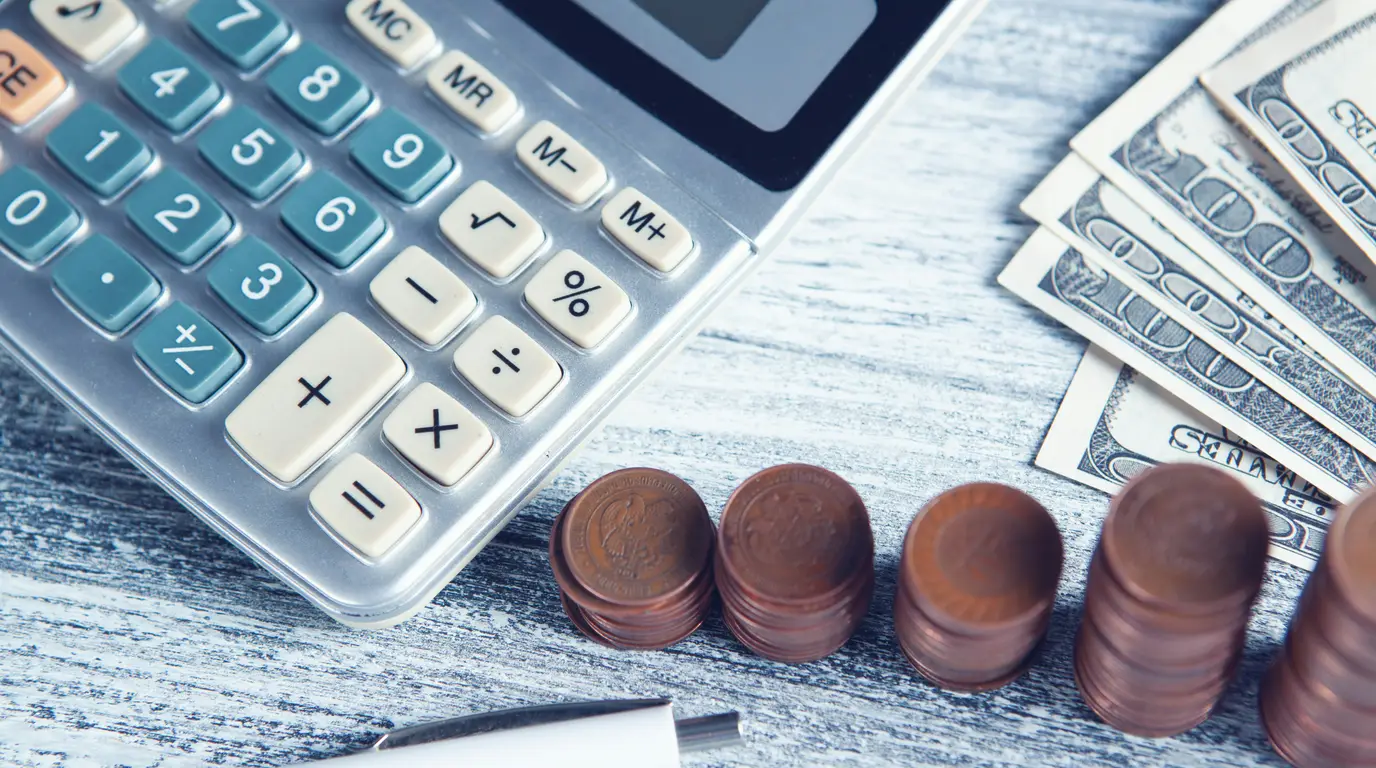 A silver calculator with teal keys beside stacks of copper coins and fanned banknotes on a pale, weathered wooden surface.