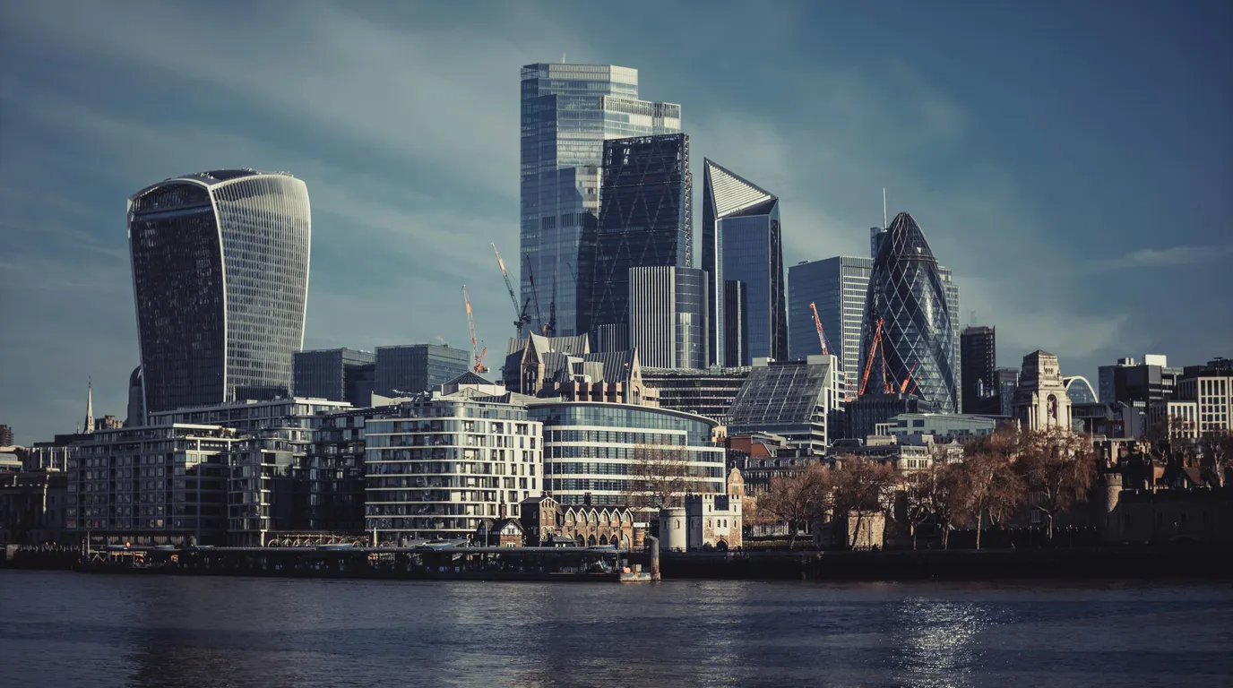 A cluster of glass and steel high-rise towers on a riverbank under a pale winter sky, with dark water in the foreground.