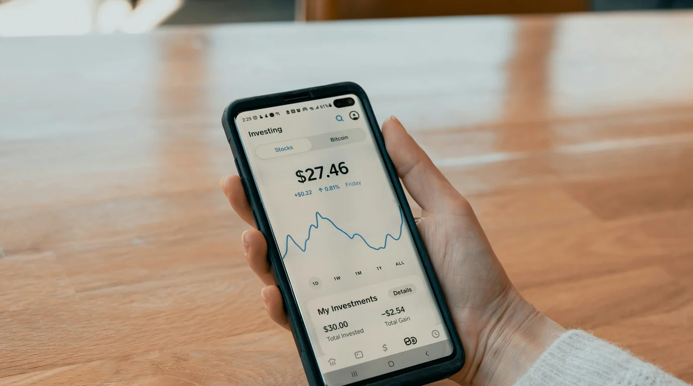 A hand holding a phone above a wooden table, its screen showing a balance, a blue line chart and investment totals.