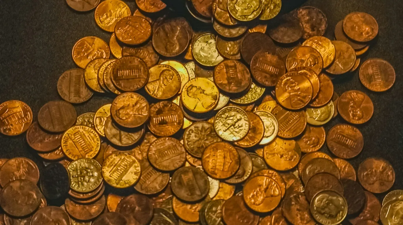 A dense pile of copper and brass coins spread across a dark surface under warm light.