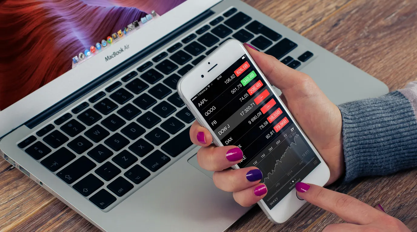 A hand with brightly painted nails holds a smartphone showing red and green market tickers above an open laptop keyboard on a wooden desk.
